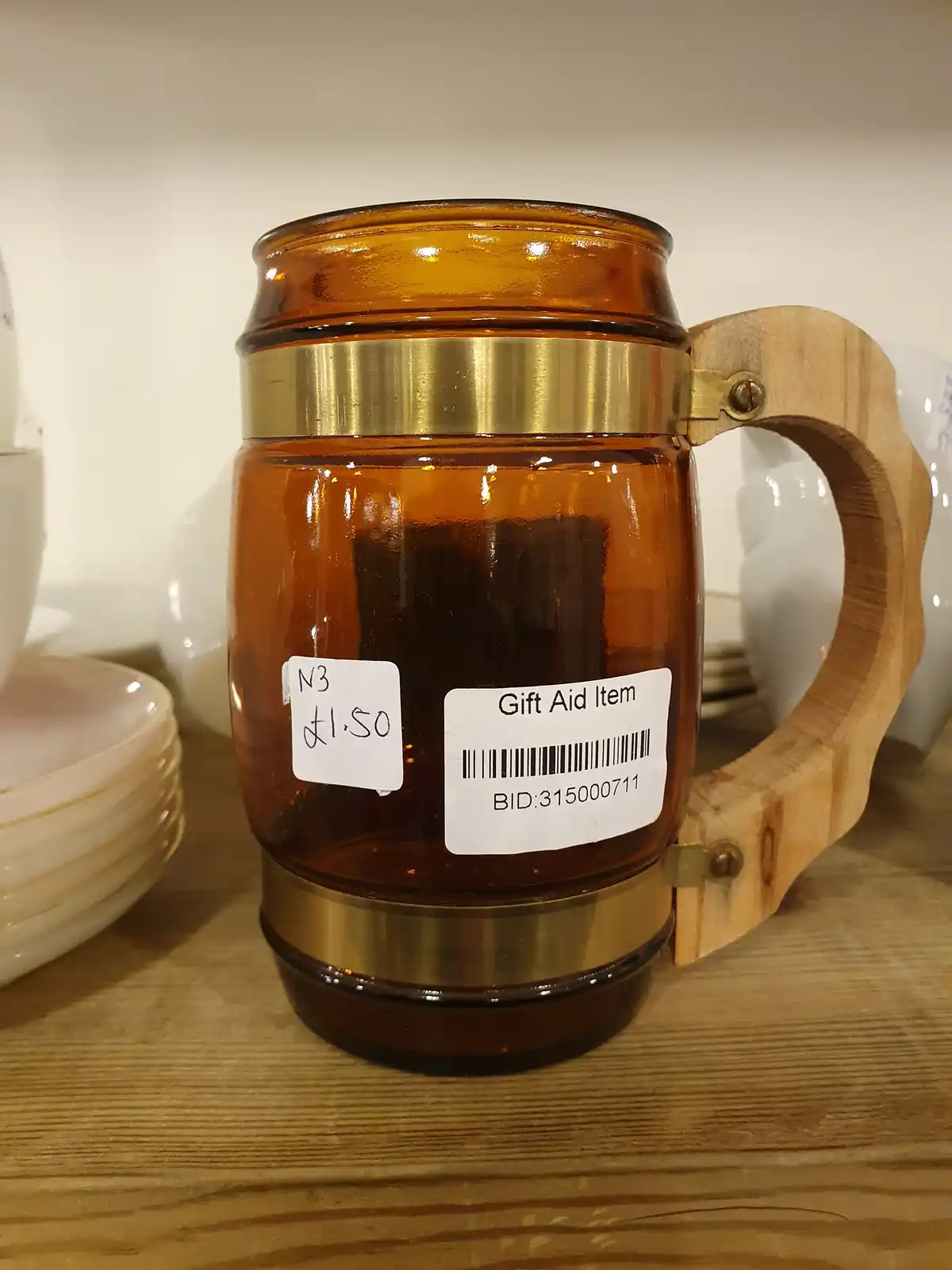 A brown glass mug with a wooden handle and brass bands sits on a wooden shelf, displaying two white labels: one handwritten…