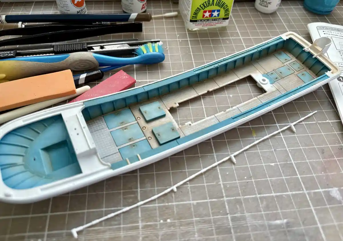 A partially assembled model boat hull sits on a gridded work mat, surrounded by modeling tools like brushes, sandpaper, and…