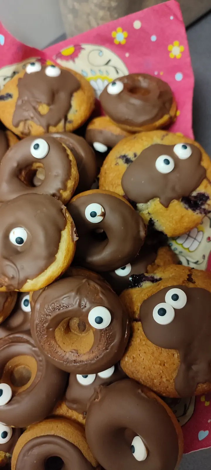 A pile of chocolate-frosted donuts and blueberry muffins, many decorated with googly eyes, resting on a pink floral napkin…