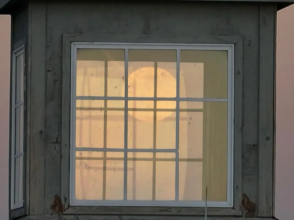 A large, round, yellowish moon is visible through a multi-paned white-framed window set in a weathered gray concrete wall…