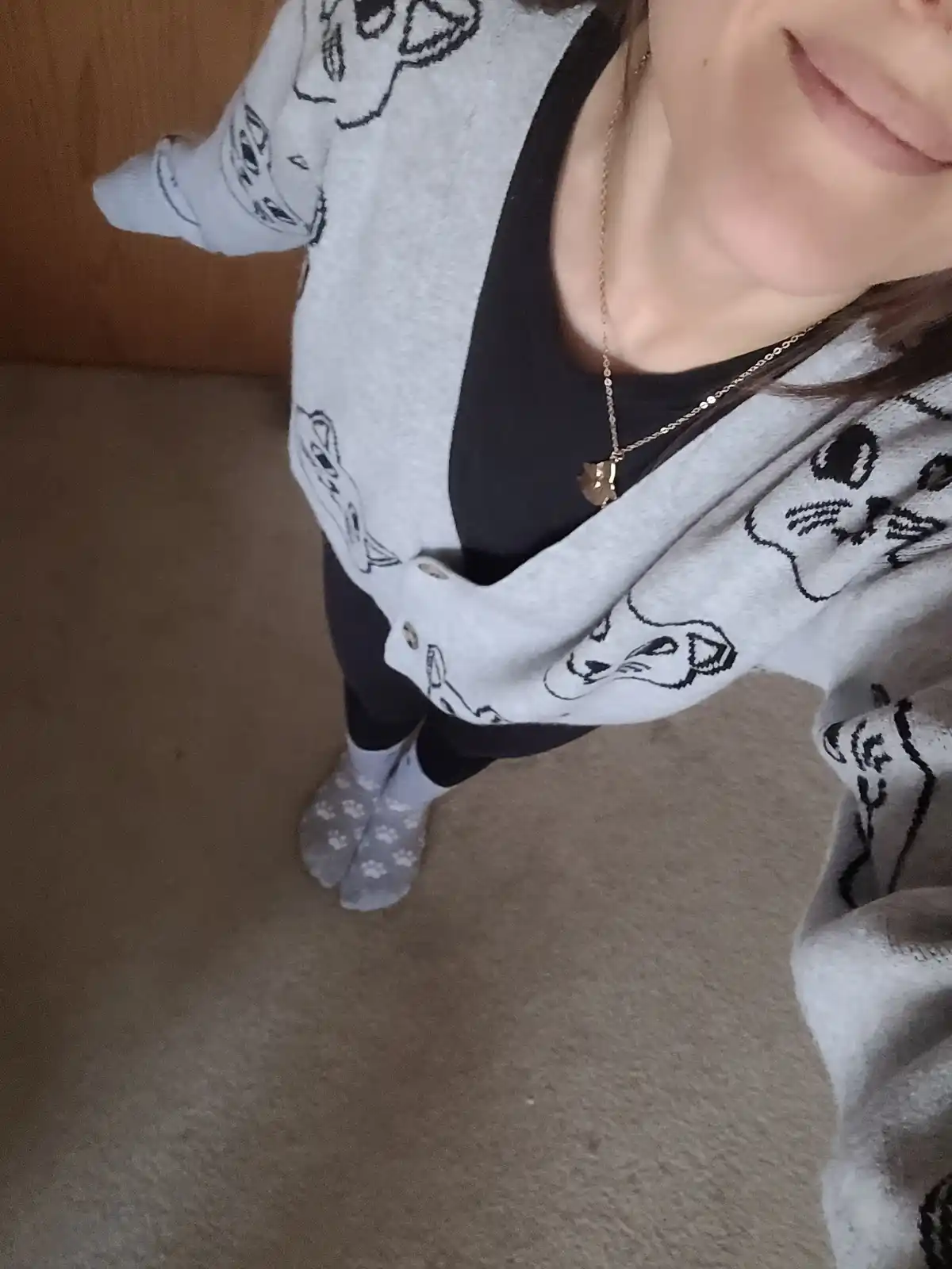 A person is shown from the chest down, wearing a grey cardigan with black cat outlines, a black top, and socks with white…