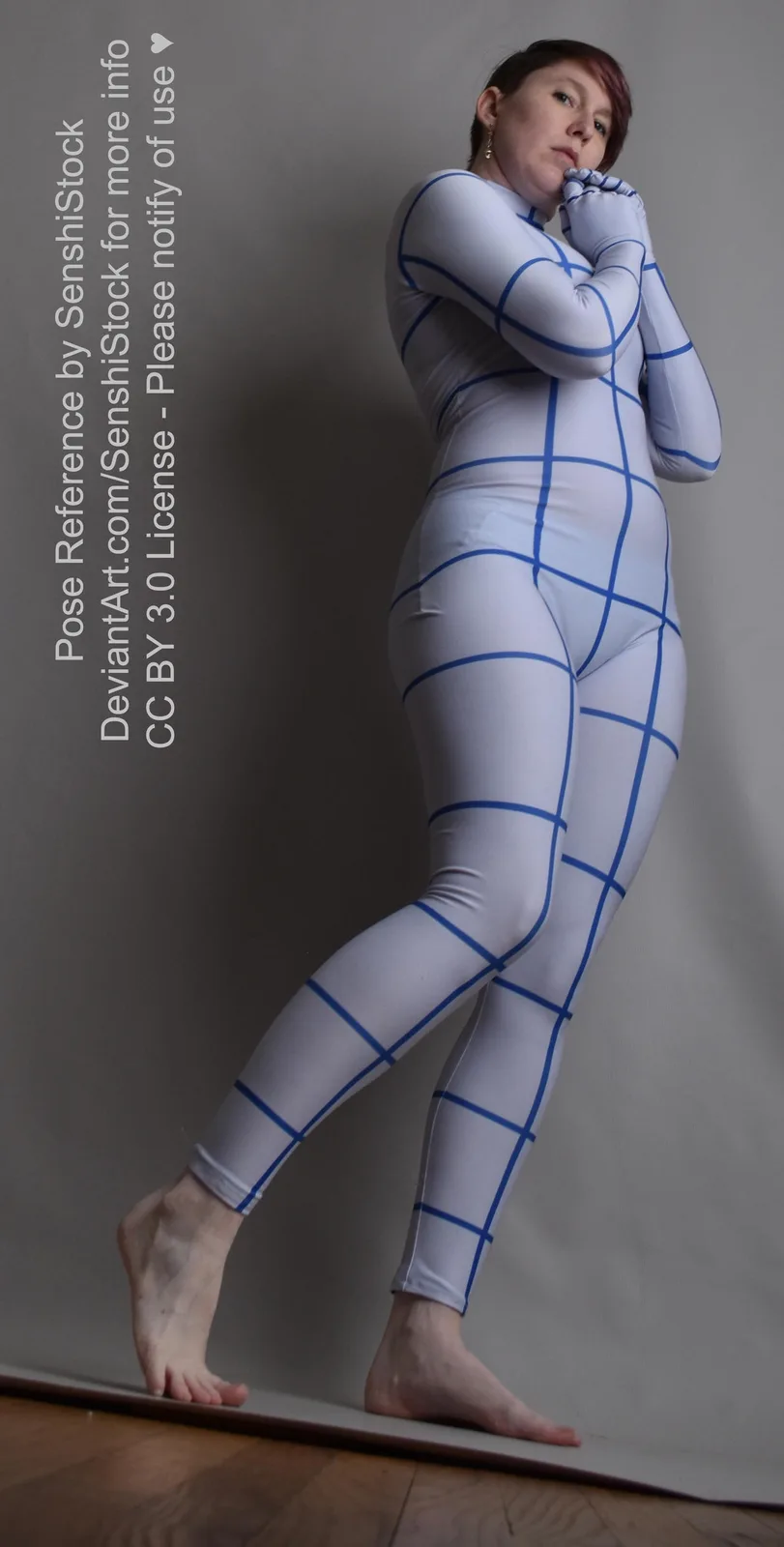 A person stands in a full-body pose wearing a white, form-fitting bodysuit with a blue grid pattern, arms crossed over their…