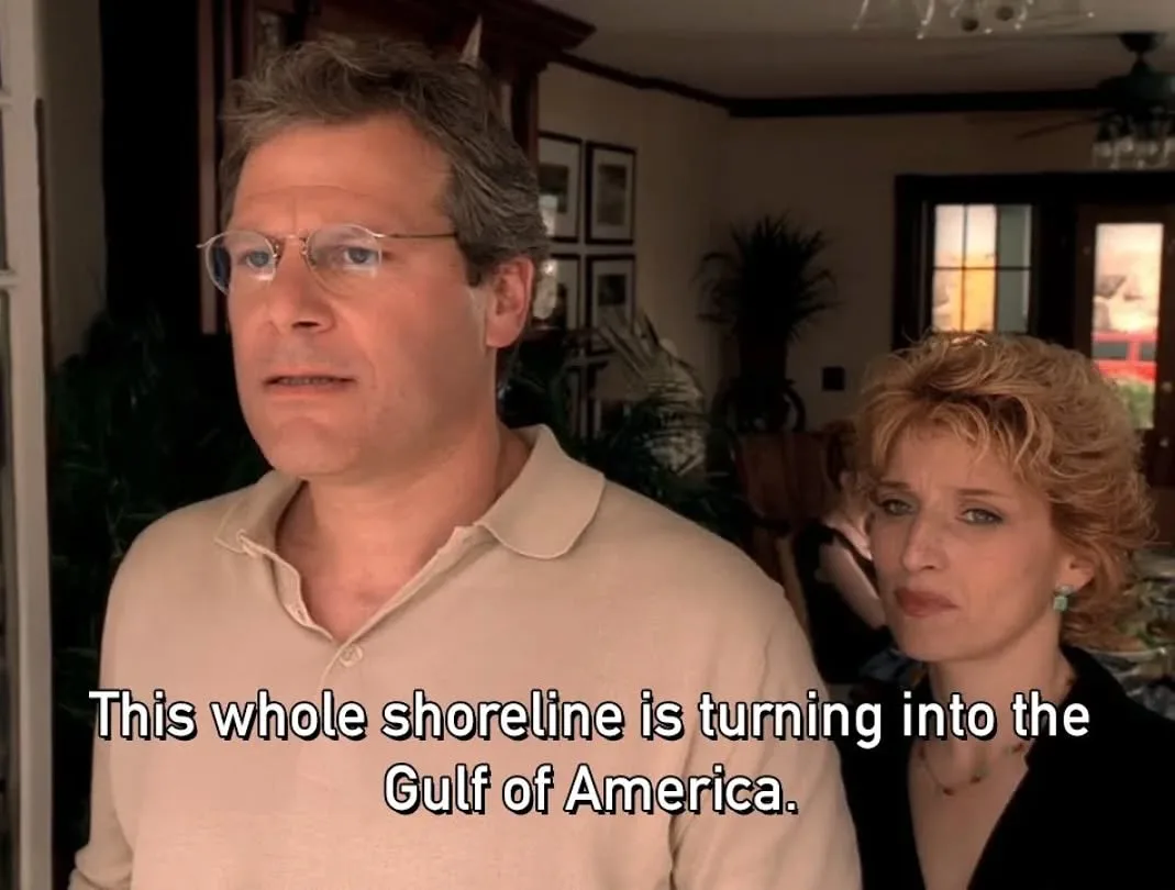A still from a TV show shows a man in glasses and a beige polo shirt looking concerned, with a woman in a black top beside…