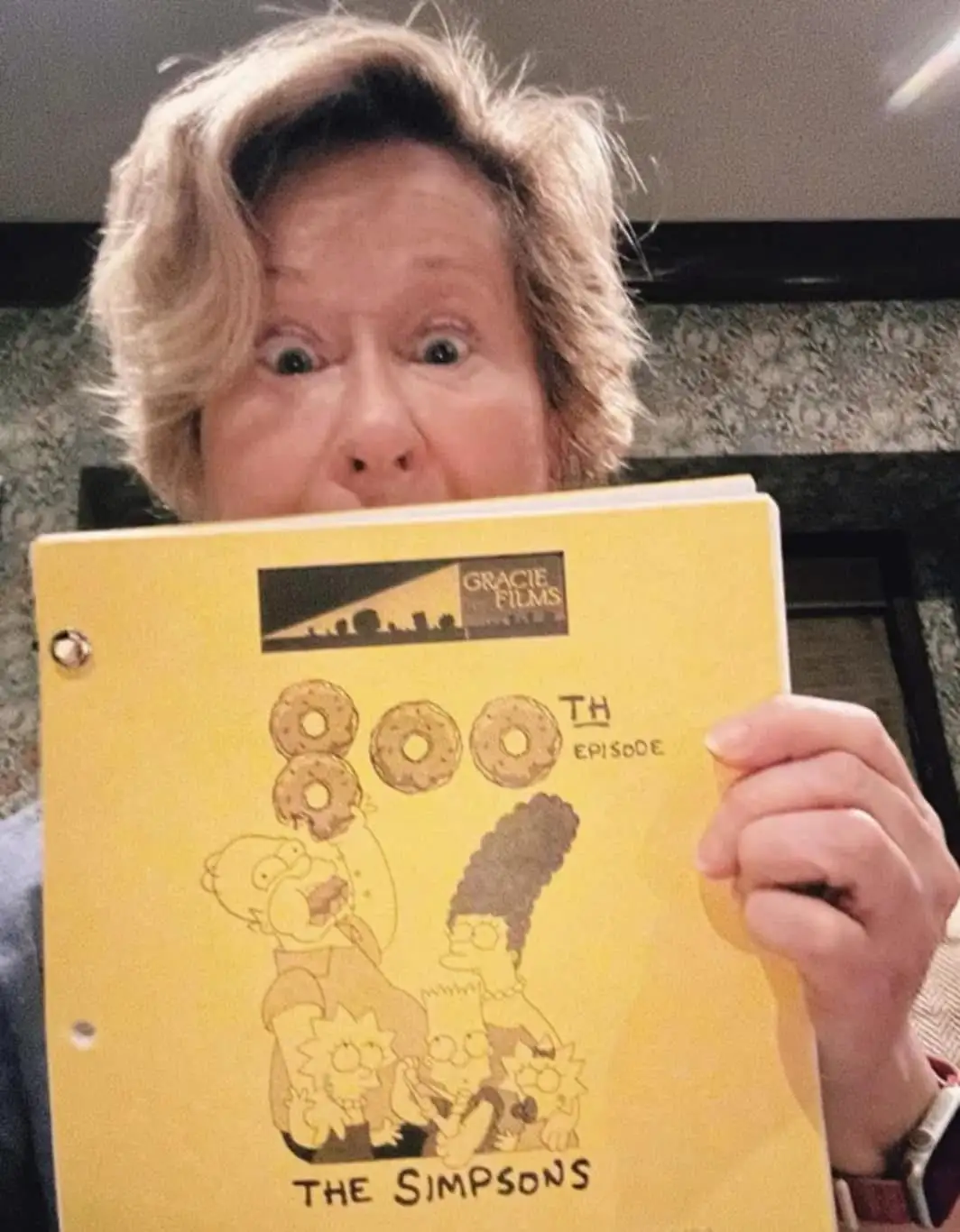 A person with wide, surprised eyes holds up a yellow 'The Simpsons' script binder. The binder cover shows the Simpson family…
