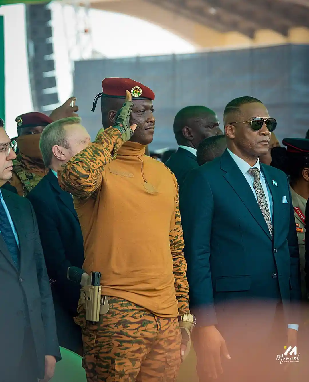 A man in a tiger-striped military uniform and red beret salutes formally while standing next to a man in a dark suit and…