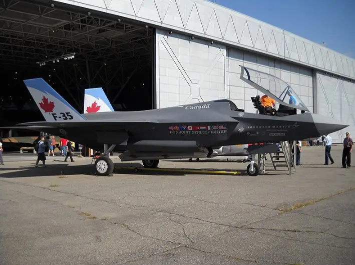 A grey F-35 Joint Strike Fighter jet is parked on tarmac outside a large hangar, its tail fins prominently displaying the…