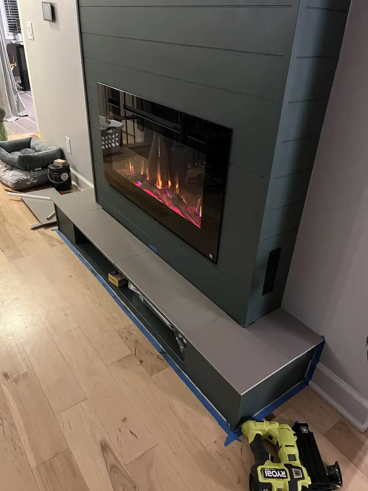 A modern electric fireplace with glowing artificial flames is set into a dark gray shiplap wall, with its matching gray…