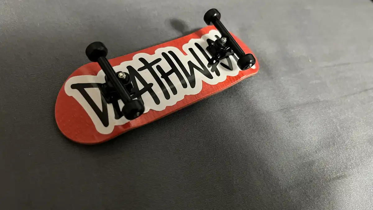 A small red fingerboard skateboard with black trucks and wheels rests on a gray fabric surface. The deck features a large…