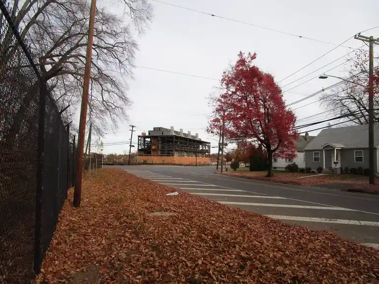 A street scene in autumn with fallen leaves covering the ground, a chain-link fence on the left, a residential house on the…