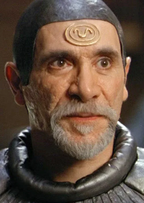 a close-up of a man with a grey beard and a black head covering, featuring a gold spiral symbol on his forehead. He wears a…