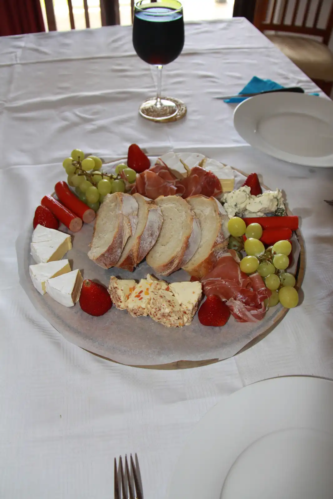 A charcuterie board with sliced bread, various cheeses, prosciutto, grapes, and strawberries is arranged on a wooden platter…