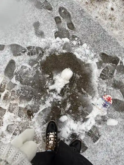 A top-down view shows a person's feet in black boots standing on a snowy, icy surface with visible footprints. In the center…