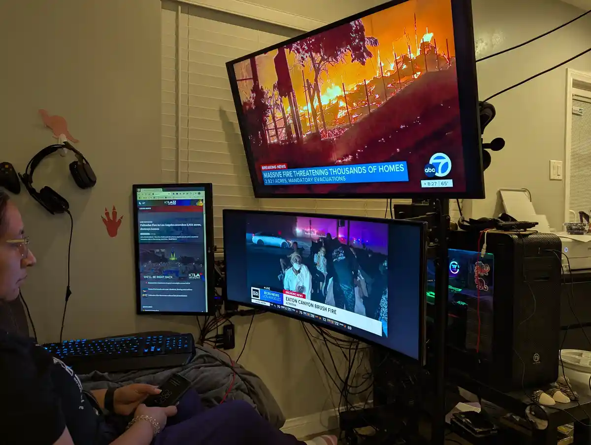 A person sits in a room with multiple monitors displaying breaking news coverage of a massive wildfire, including footage of…