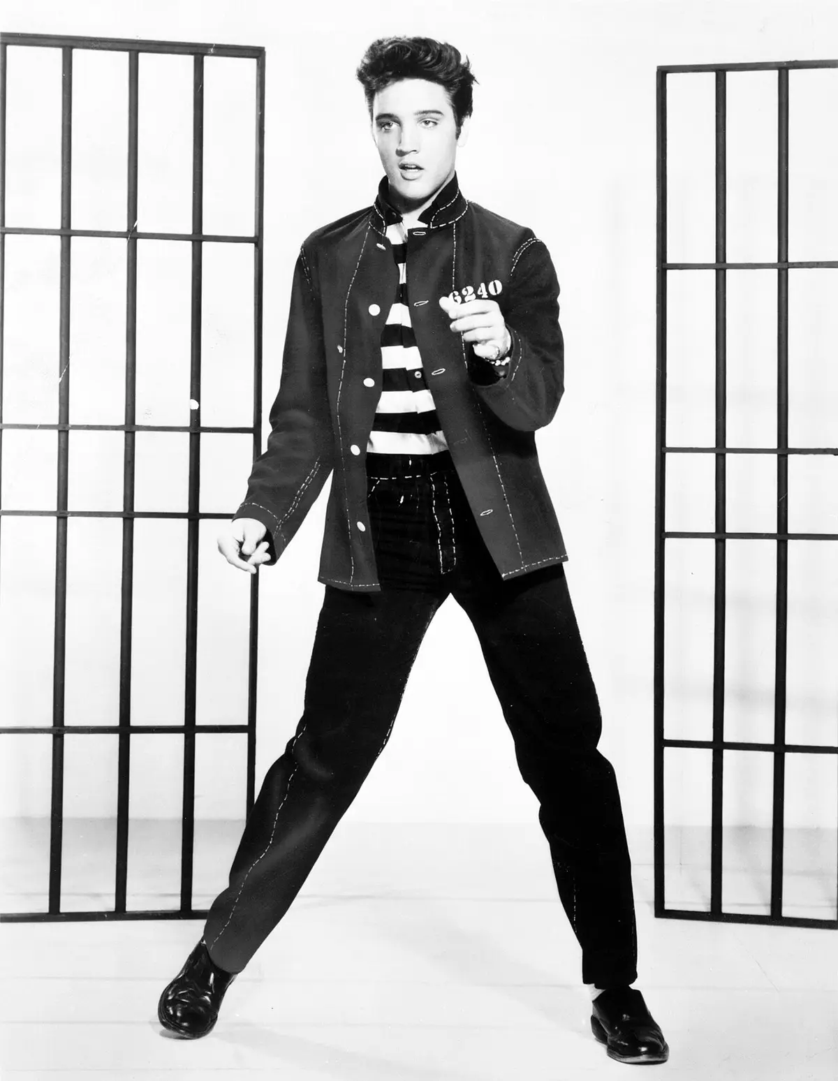A black-and-white photo of Elvis Presley posing between two sets of prison bars, wearing a striped prison jumpsuit under a…