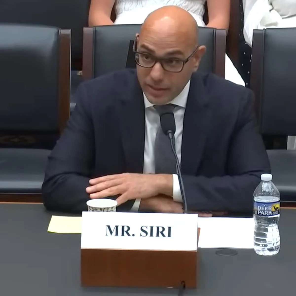 A bald man in a dark suit and glasses sits at a formal table with a microphone, a nameplate reading 'MR. SIRI', a paper cup…