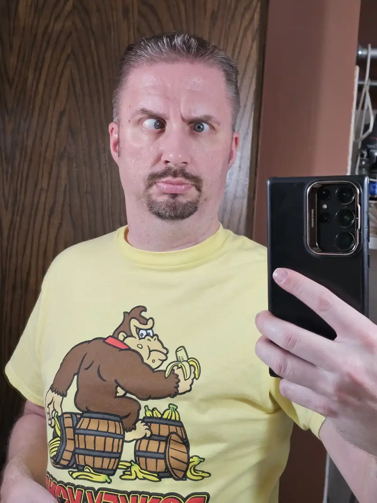 A man with a goatee and short hair takes a selfie in a mirror while wearing a yellow t-shirt featuring the character Donkey…