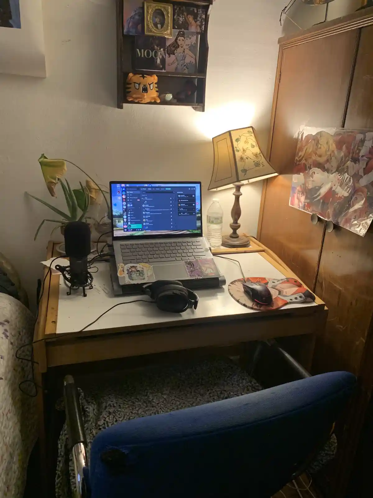 A cozy, cluttered desk setup with a laptop displaying a Discord interface, a microphone, headphones, and anime posters on…