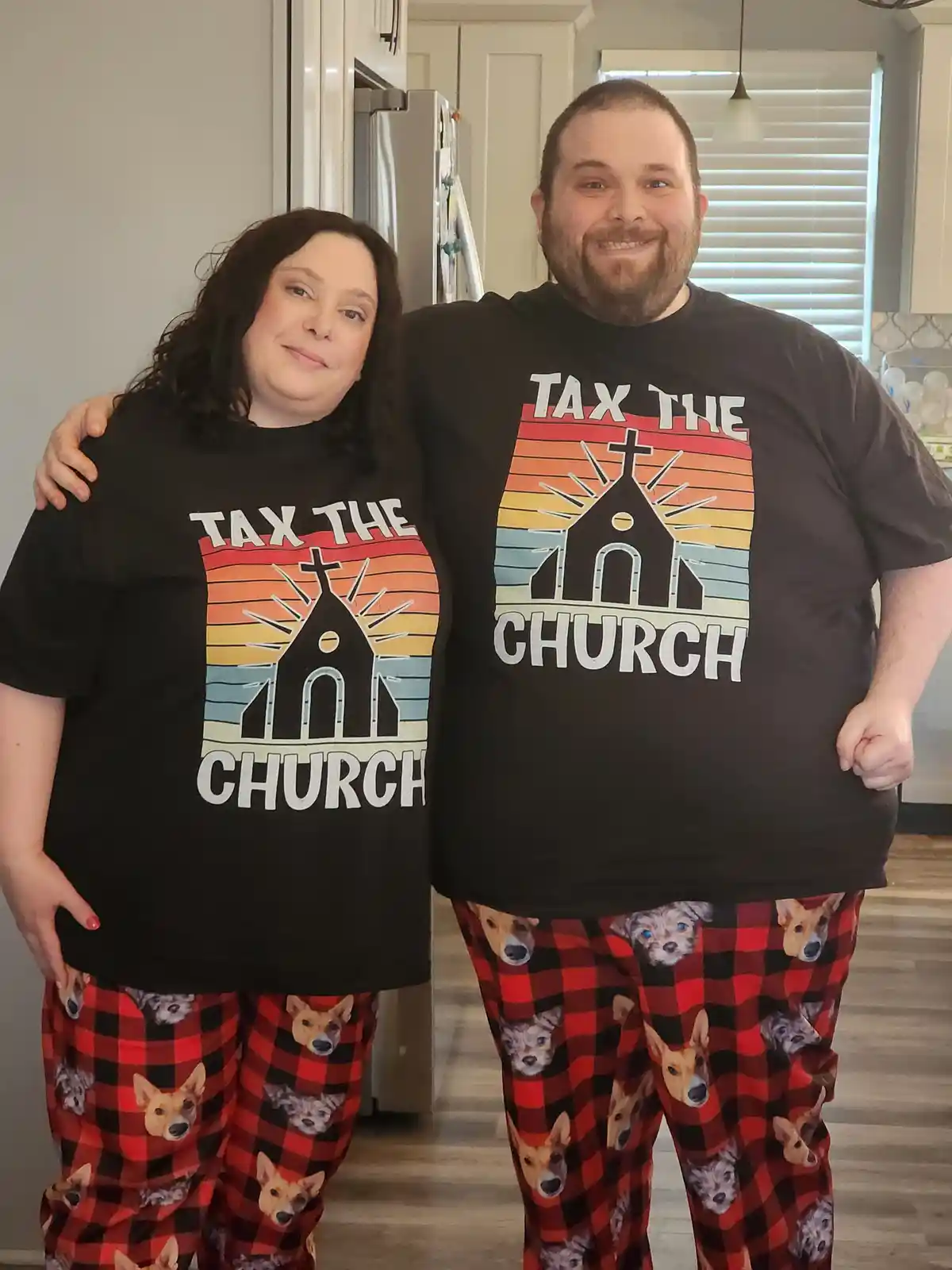 A man and woman stand together in a kitchen, both wearing matching black t-shirts with the text 'TAX THE CHURCH' over a…