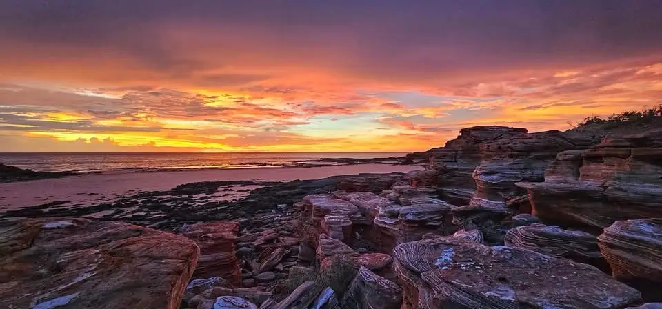 A vibrant sunset over a rocky beach with layered red and orange cliffs in the foreground, the sky ablaze with fiery hues…