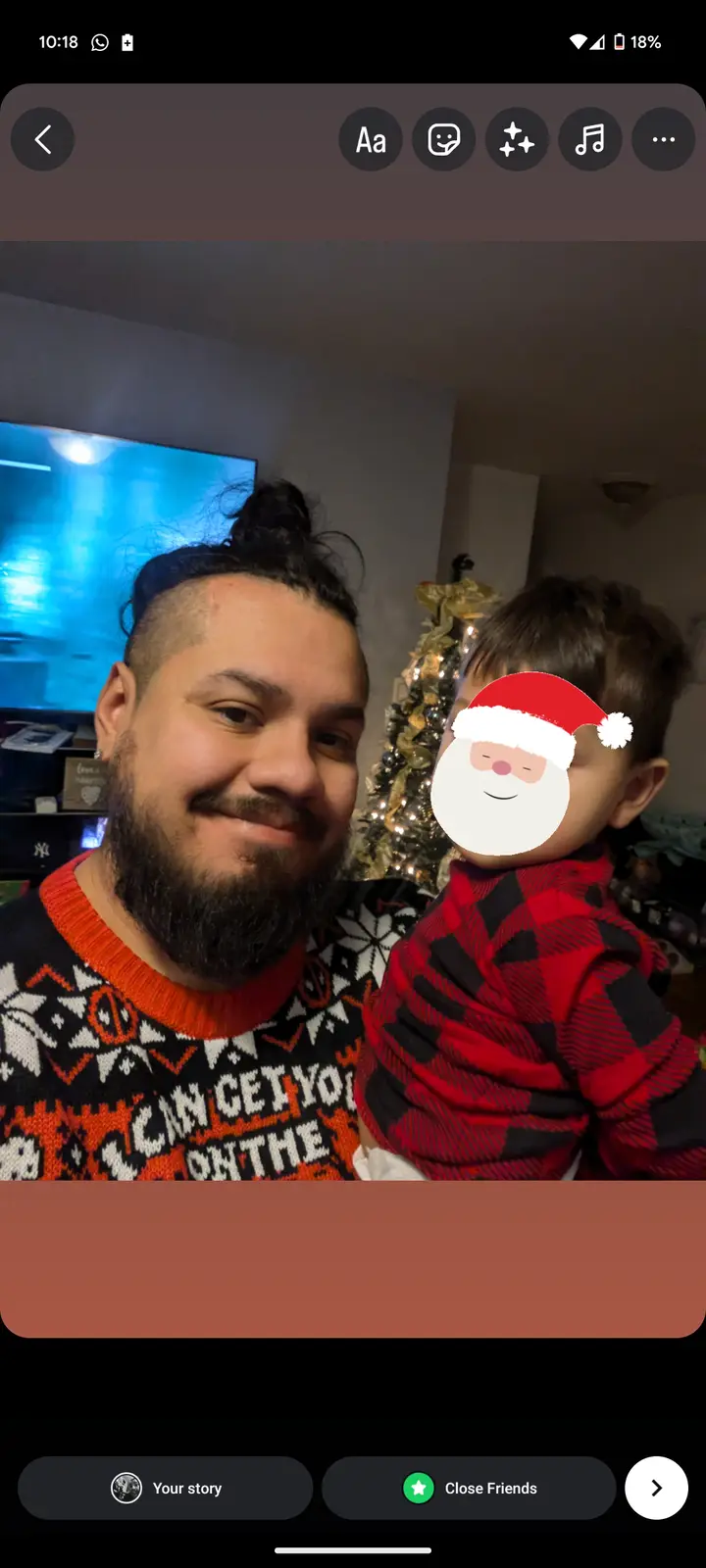 A man with a beard and festive sweater holds a child wearing a red plaid shirt; the child's face is obscured by a cartoon…