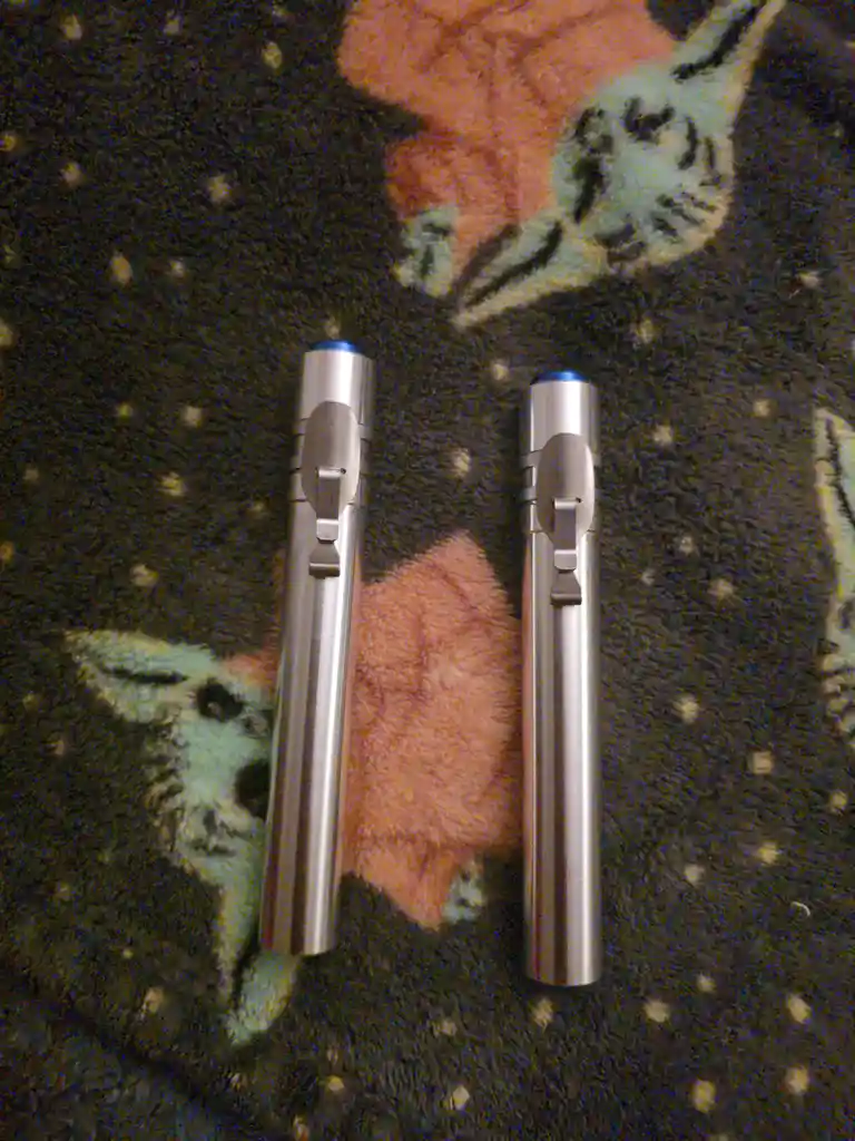 Two identical silver cylindrical pens with blue caps lie side by side on a dark fabric with a cat pattern. The overlaid text…