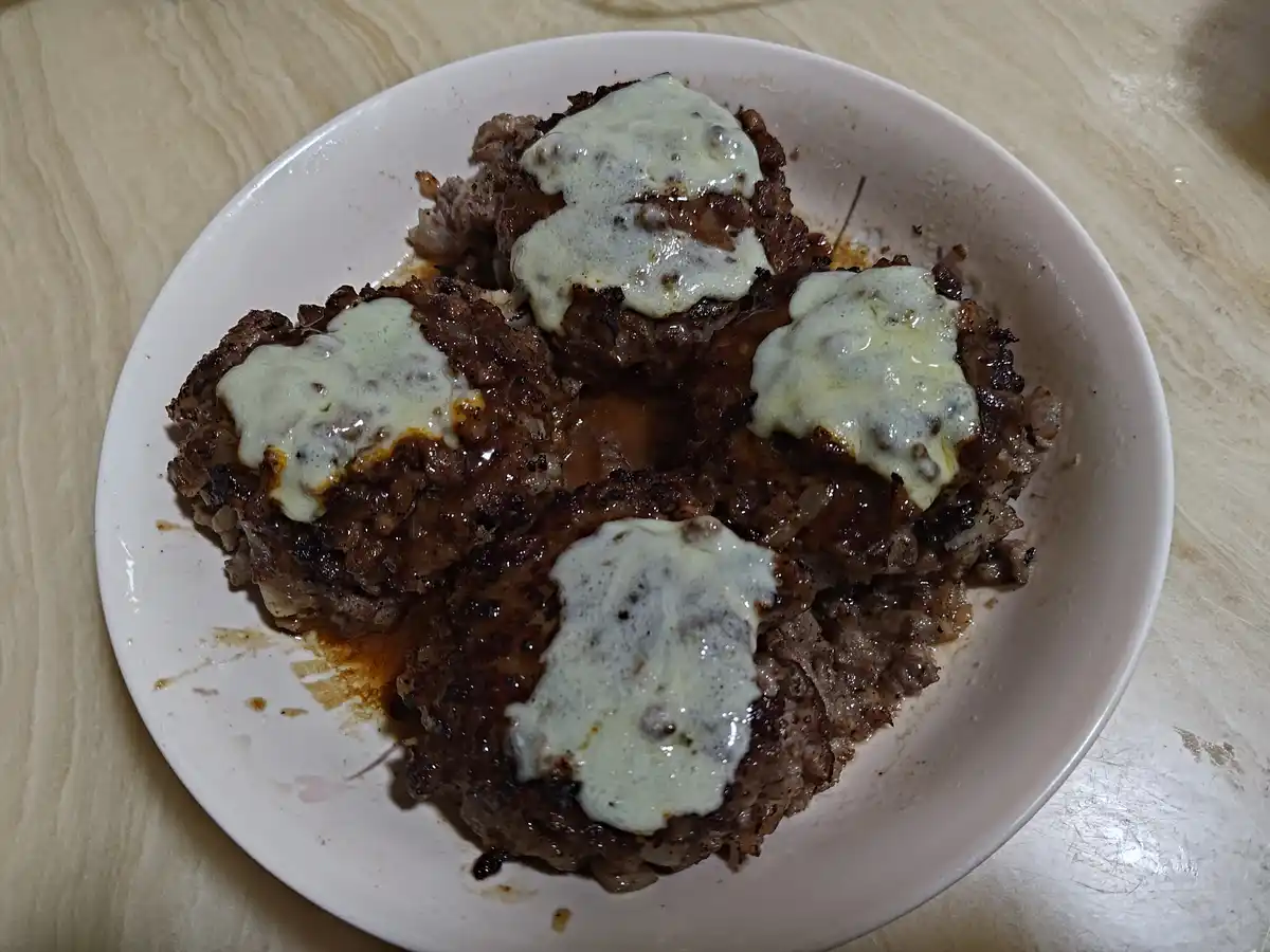 A white plate holds four browned, round meat patties, each topped with a dollop of melted white cheese, sitting on a…