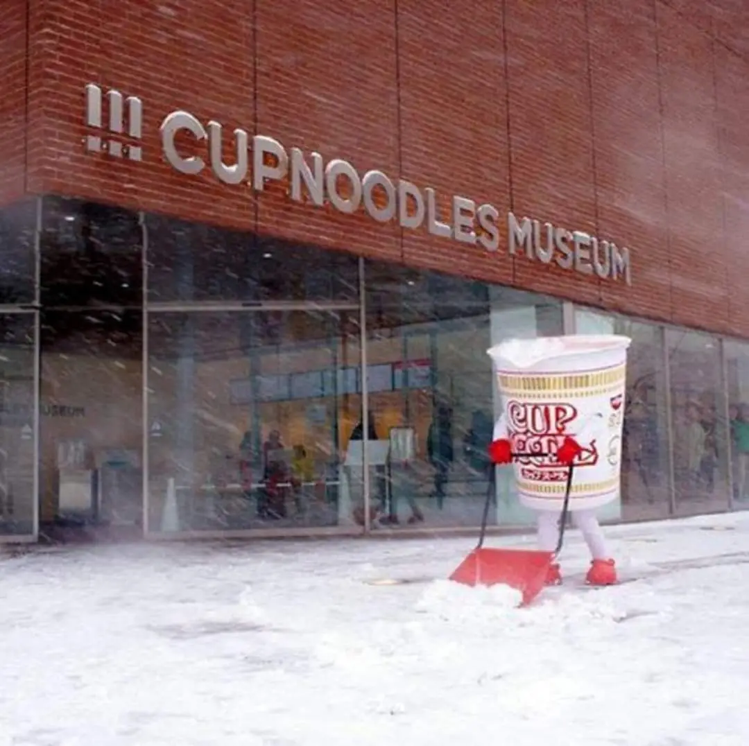 A person in a full-sized Cup Noodles cup costume is shoveling snow outside the Cup Noodles Museum, which has large silver…