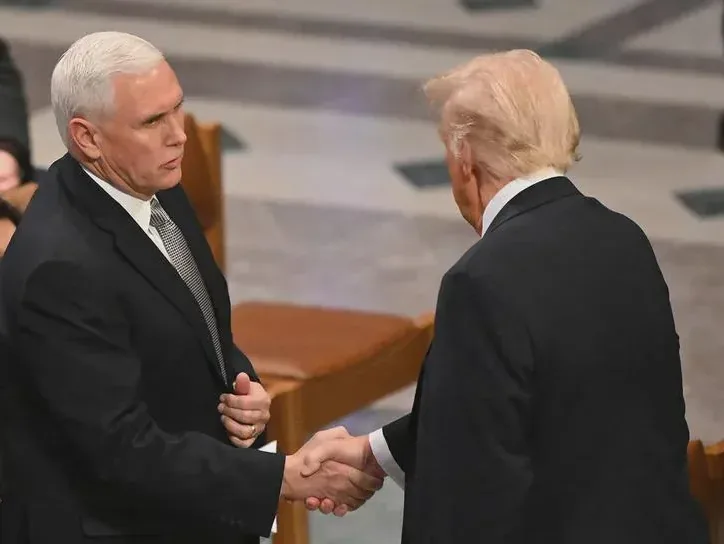 Two men in dark suits shake hands in a formal setting. The man on the left, with white hair and a stern expression, looks…