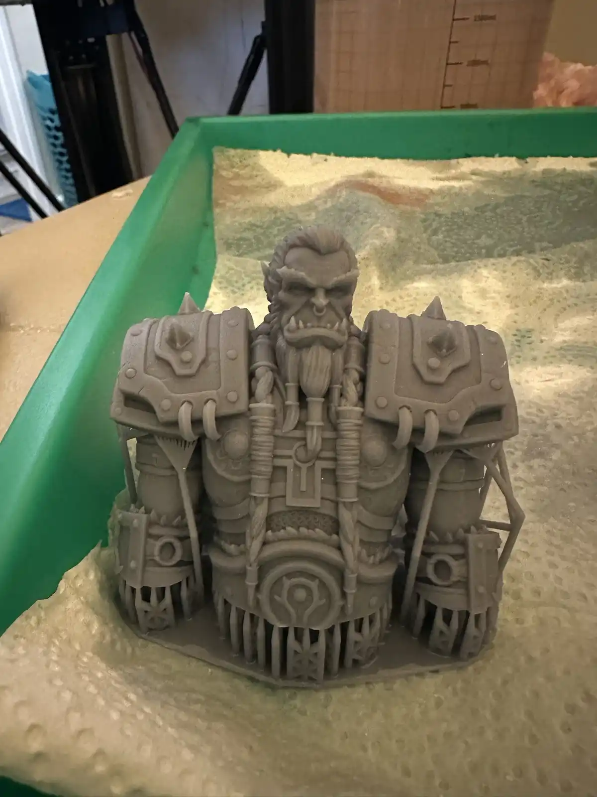 A gray, un-painted 3D-printed figurine of a fantasy orc warrior with a beard and spiked armor stands on a textured base…