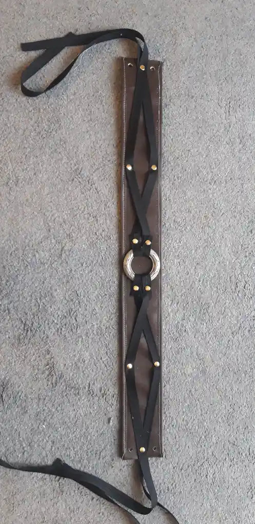 A handmade leather harness with black straps and gold-colored rivets lies on a gray carpet. It features a central silver…