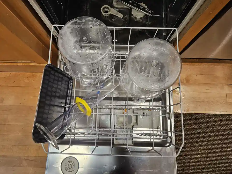 An overhead shot shows a dishwasher rack with two large, clear blender jars upside down, a clear measuring cylinder with a…