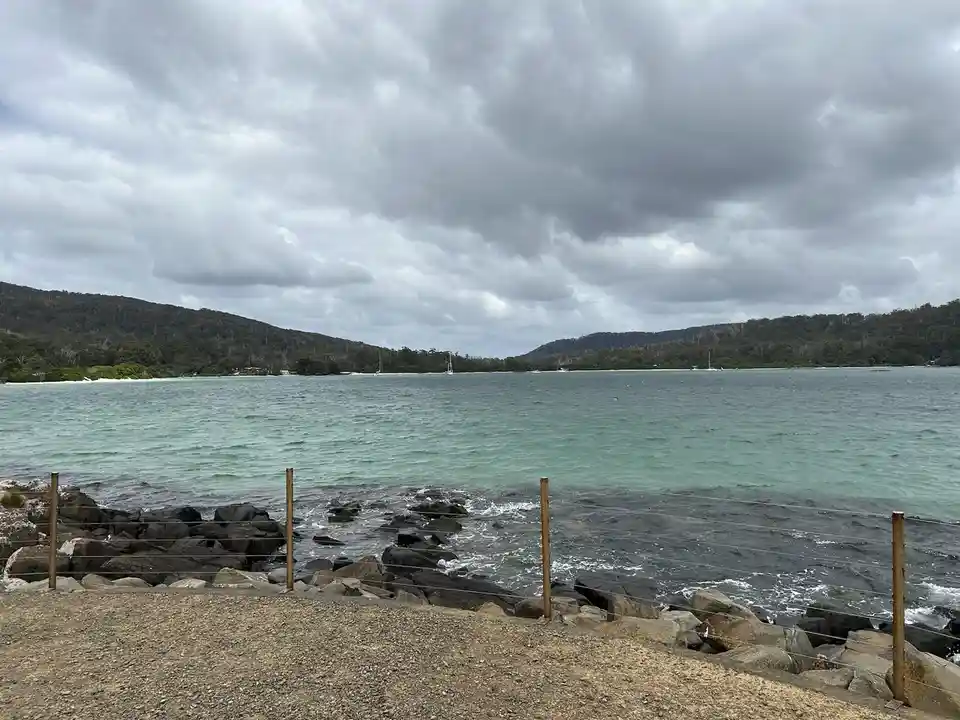 A scenic photo of a calm bay with turquoise water, distant forested hills, and a cloudy sky, framed by a gravel path and a…