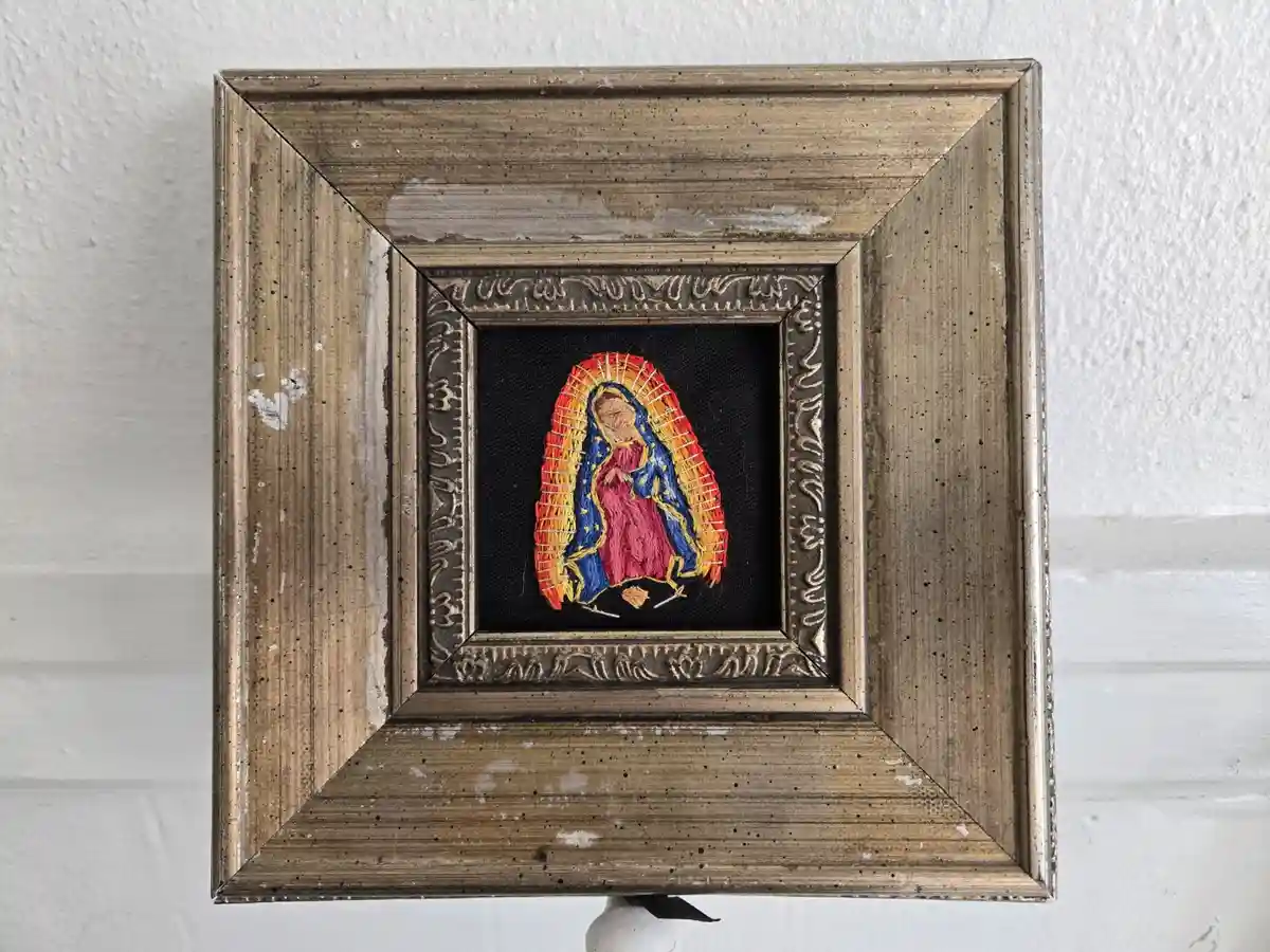 A small, framed embroidered image of the Virgin of Guadalupe is mounted on a white wall. The embroidery shows her in…