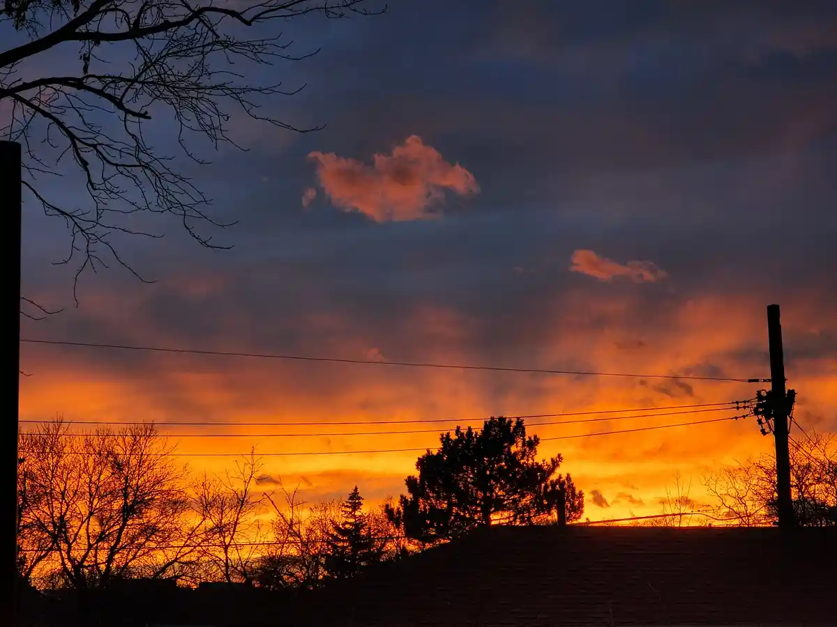 A dramatic sunset with fiery orange and purple clouds silhouetting bare trees, power lines, and a roof. The overlaid text…