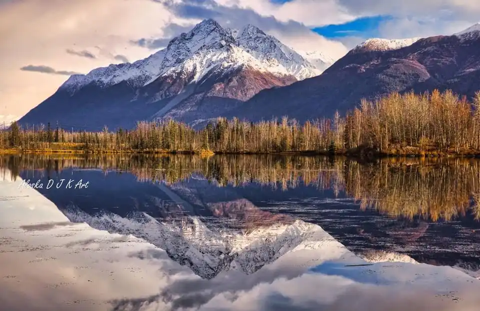 A serene landscape photo shows snow-capped mountains reflected perfectly in a calm lake, with autumn-colored trees along the…
