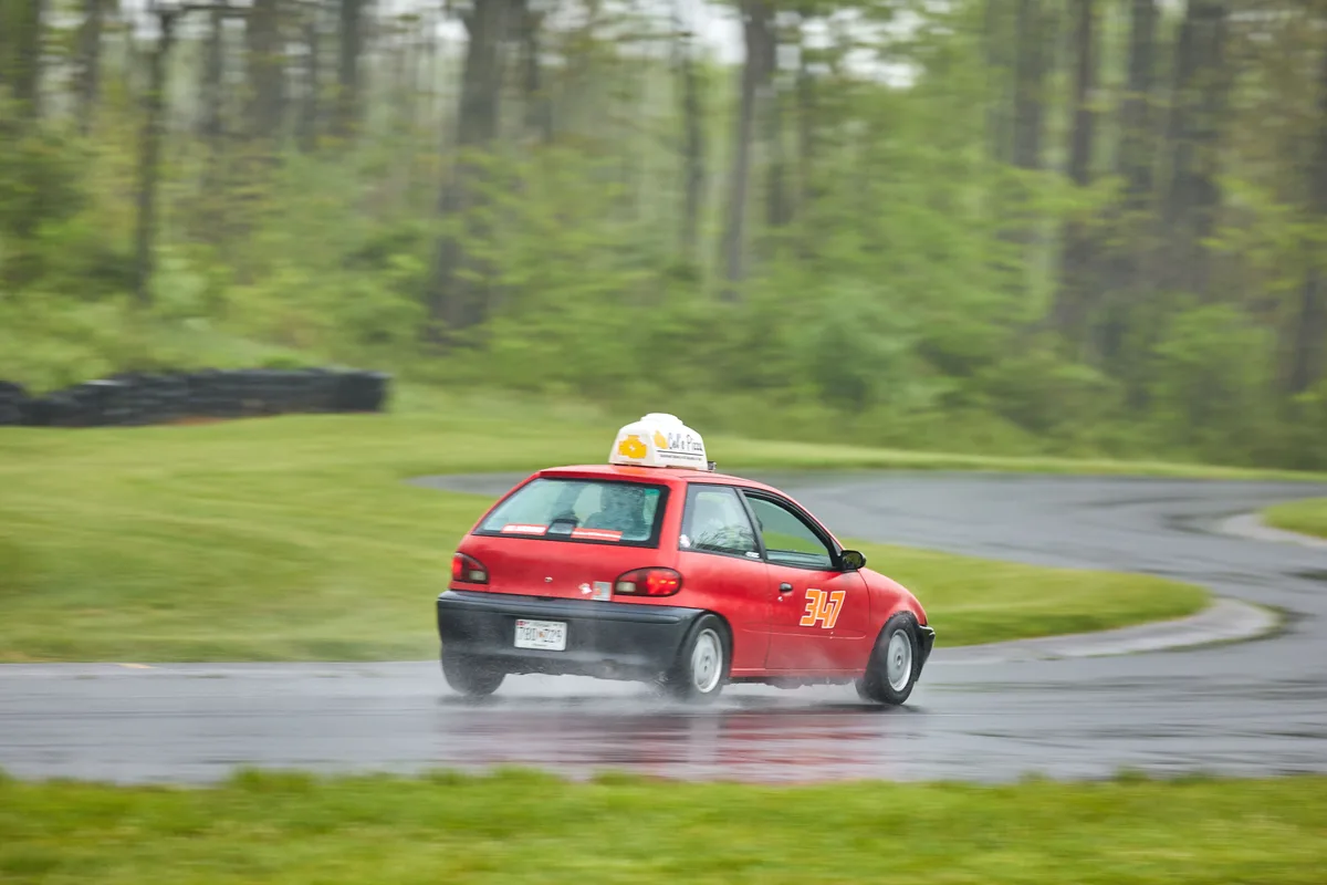 A red compact car with a 'Call e Pizza' sign on its roof is speeding around a wet, curving road in a forested area, kicking…
