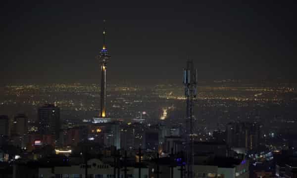 A nighttime cityscape featuring a tall, illuminated tower and a smaller communications tower in the foreground, with a…