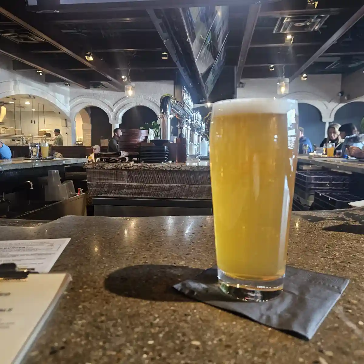 A tall glass of golden beer with a frothy head sits on a dark napkin on a speckled bar counter. In the background, a busy…