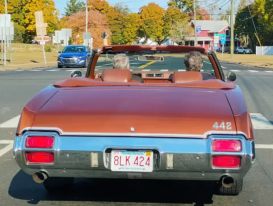 A rear view of a vintage two-tone brown and blue convertible with '442' on the trunk, showing two people driving through a…
