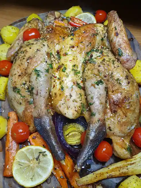 A roasted whole chicken sits on a baking tray surrounded by roasted vegetables including carrots, potatoes, cherry tomatoes…