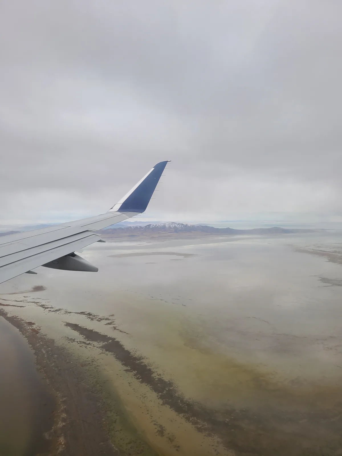 An airplane wing is visible from a passenger's window, flying over a vast, flat, salt-encrusted lake with distant…