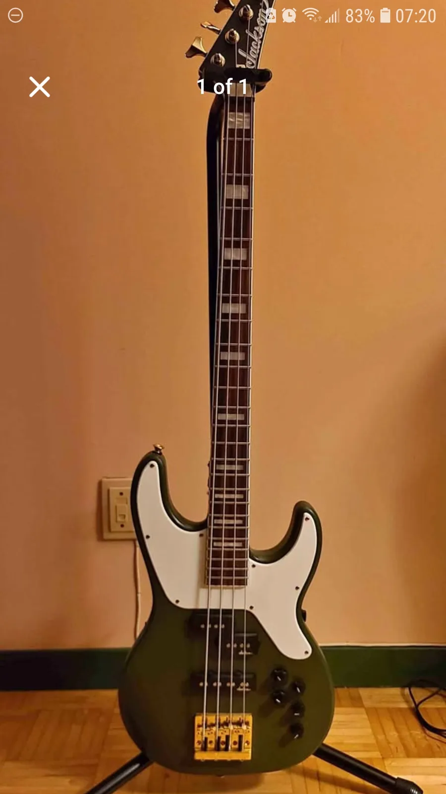 A green Jackson bass guitar with a white pickguard stands upright on a stand against a plain tan wall, with a light switch…