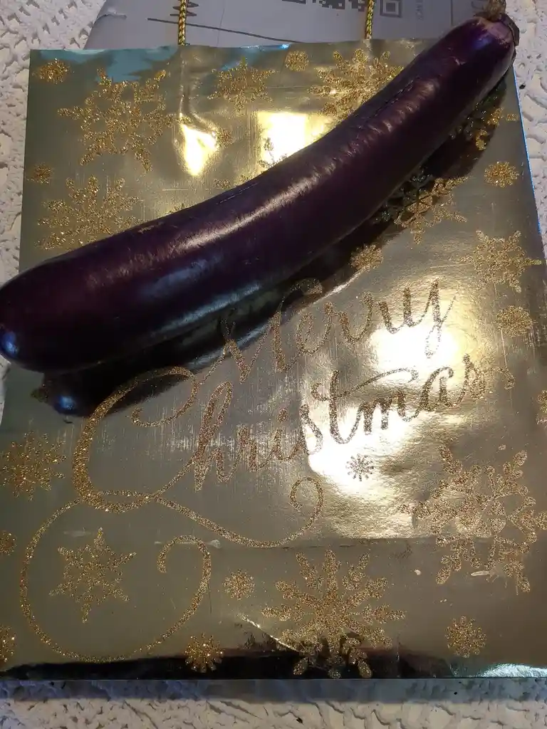 A shiny purple eggplant is placed diagonally across a gold-colored Christmas gift bag decorated with glittery snowflakes and…