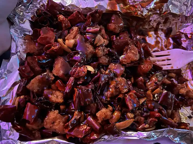 A close-up photo shows a nearly empty foil-lined container filled with spicy-looking food, likely Kung Pao chicken or a…