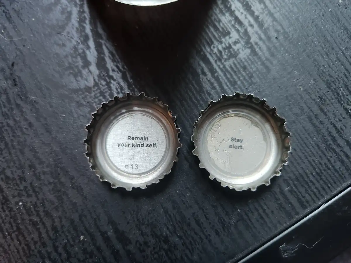 Two metal bottle caps sit side by side on a dark, textured surface. The cap on the left reads 'Remain your kind self.' and…