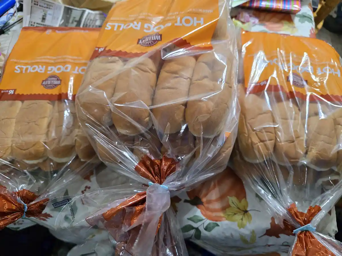 Several packages of Weisley brand hot dog rolls are piled together on a table with a fall-themed tablecloth featuring…