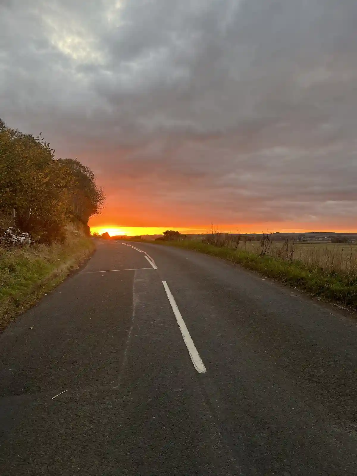 A scenic rural road stretches toward a vibrant orange and red sunset under a cloudy sky, with grassy fields and trees on…