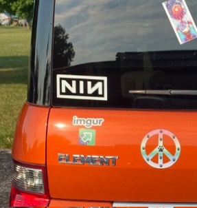 The rear of an orange car shows a black rectangular sticker with the white letters 'NIN' on the back window, alongside other…