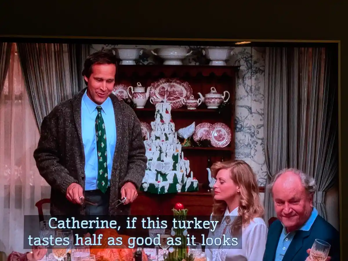 A still from a TV show shows a man in a cardigan and tie standing at a dining table with a large turkey, smiling as he…