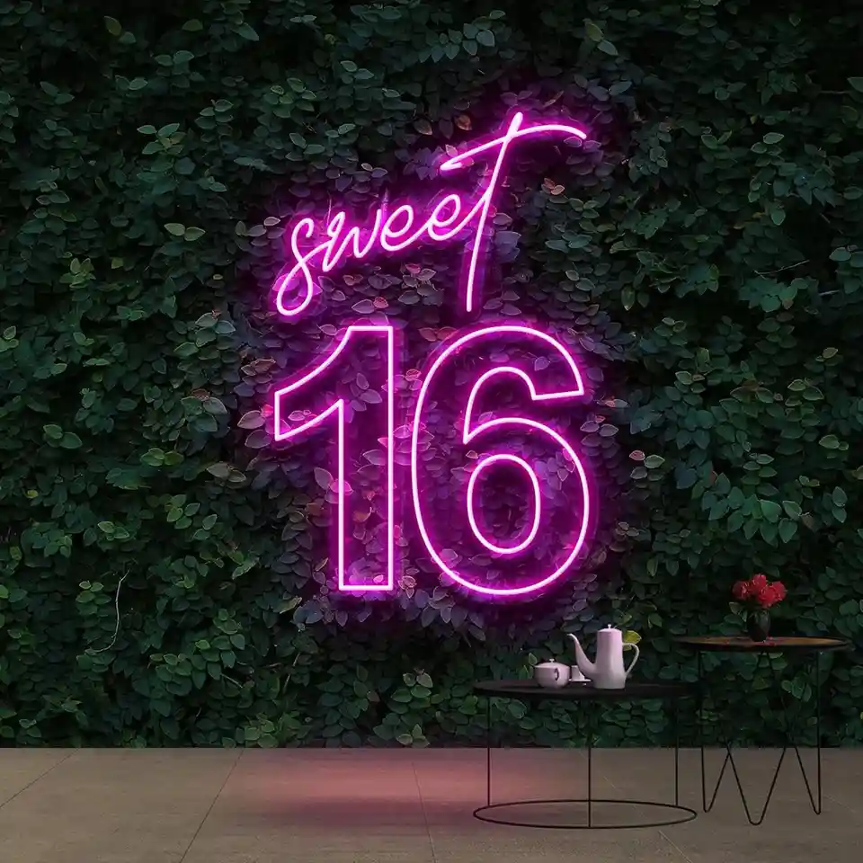A vibrant pink neon sign reading 'sweet 16' in cursive and block letters glows against a dense green leafy wall. Below, two…